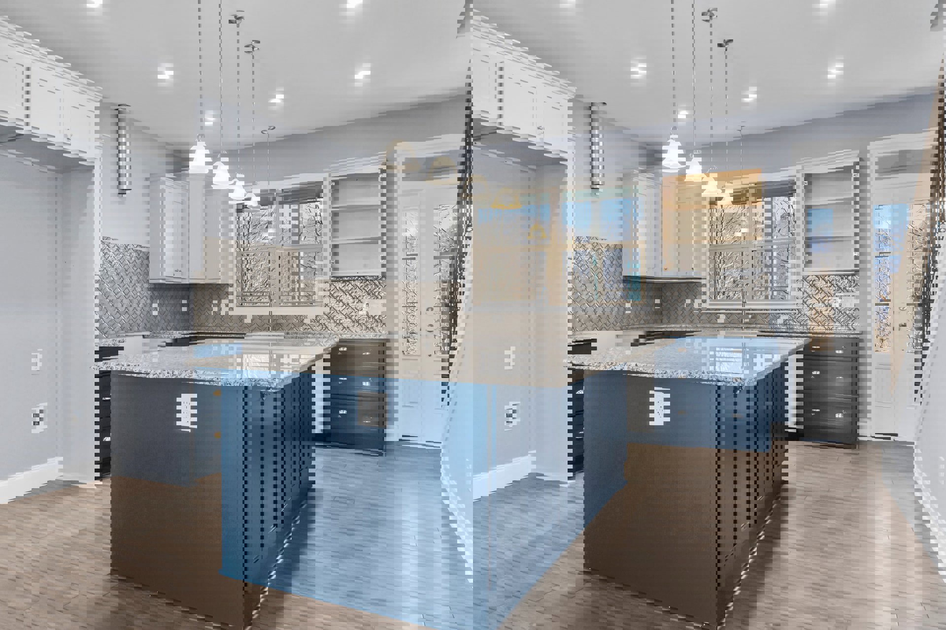 Spacious kitchen featuring a large marble-topped island, blue drawers with silver handles, white cabinetry with open shelves, tile backsplash, and a white outlet on a blue accent