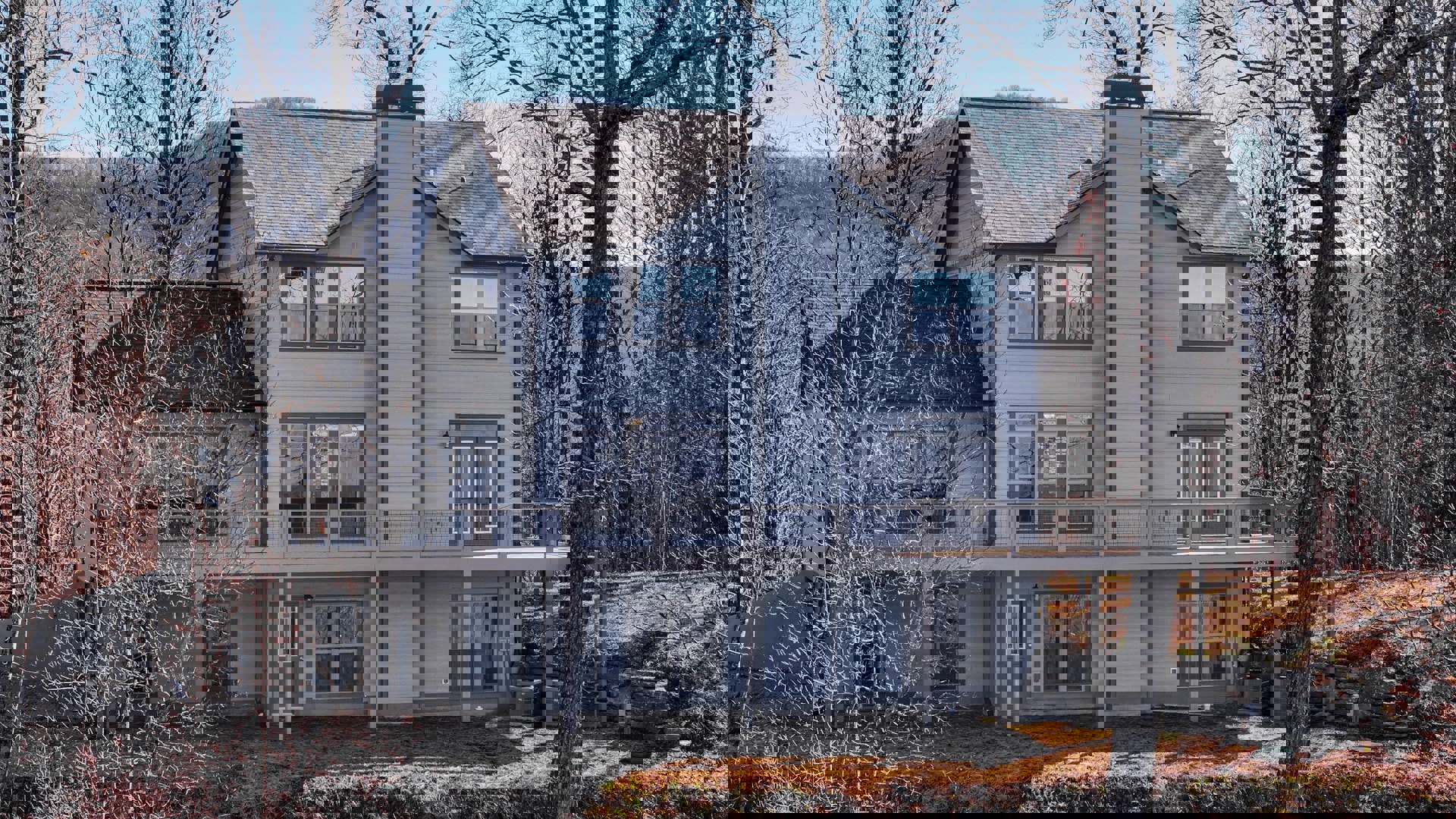 Two-story house with stone and wood siding, white-framed windows, spacious elevated deck, surrounded by mature trees and landscaped yard