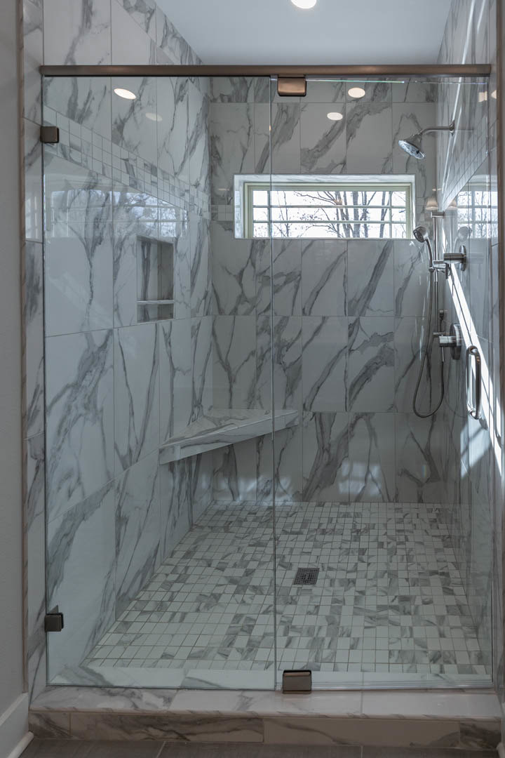 White marble shower with glass door, built-in bench, silver handle, and window overlooking leafy tree