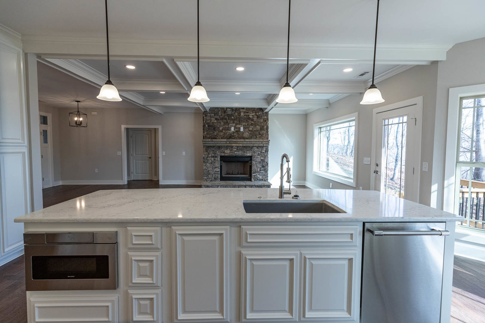 White kitchen island with built-in sink, stone fireplace with textured surround, white cabinetry, stainless steel microwave, black door handle, and multi-light chandelier hanging