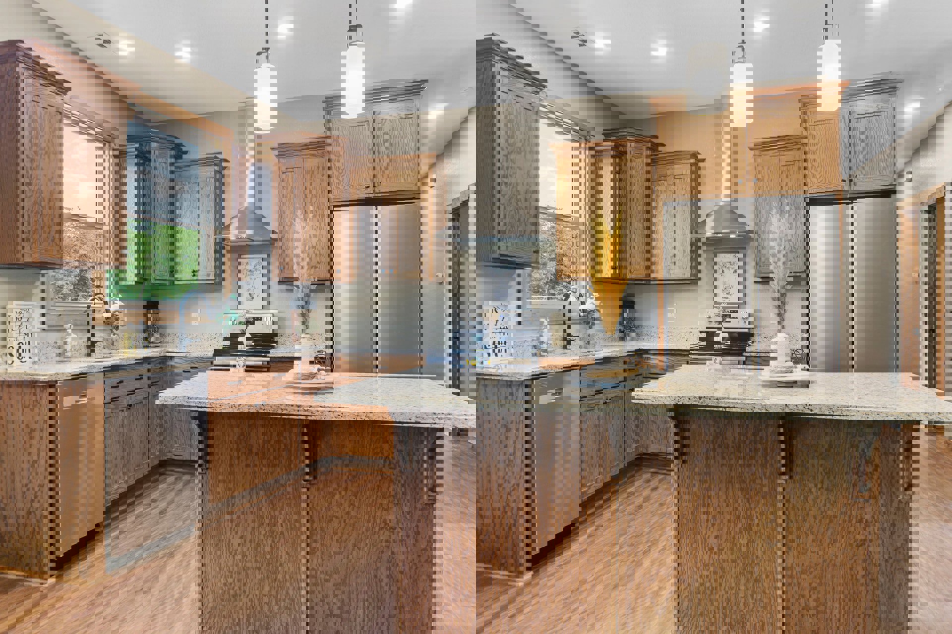 Kitchen with stainless steel refrigerator, large central island with light stone countertop, wood cabinetry, white vase with dried flowers, candle on plate, and decorative sign on