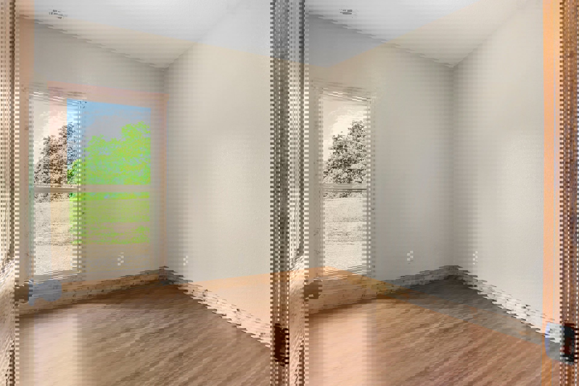 Sunlit room featuring wide wood plank flooring, large window overlooking trees and grass, white walls, and a black door knob visible in the foreground