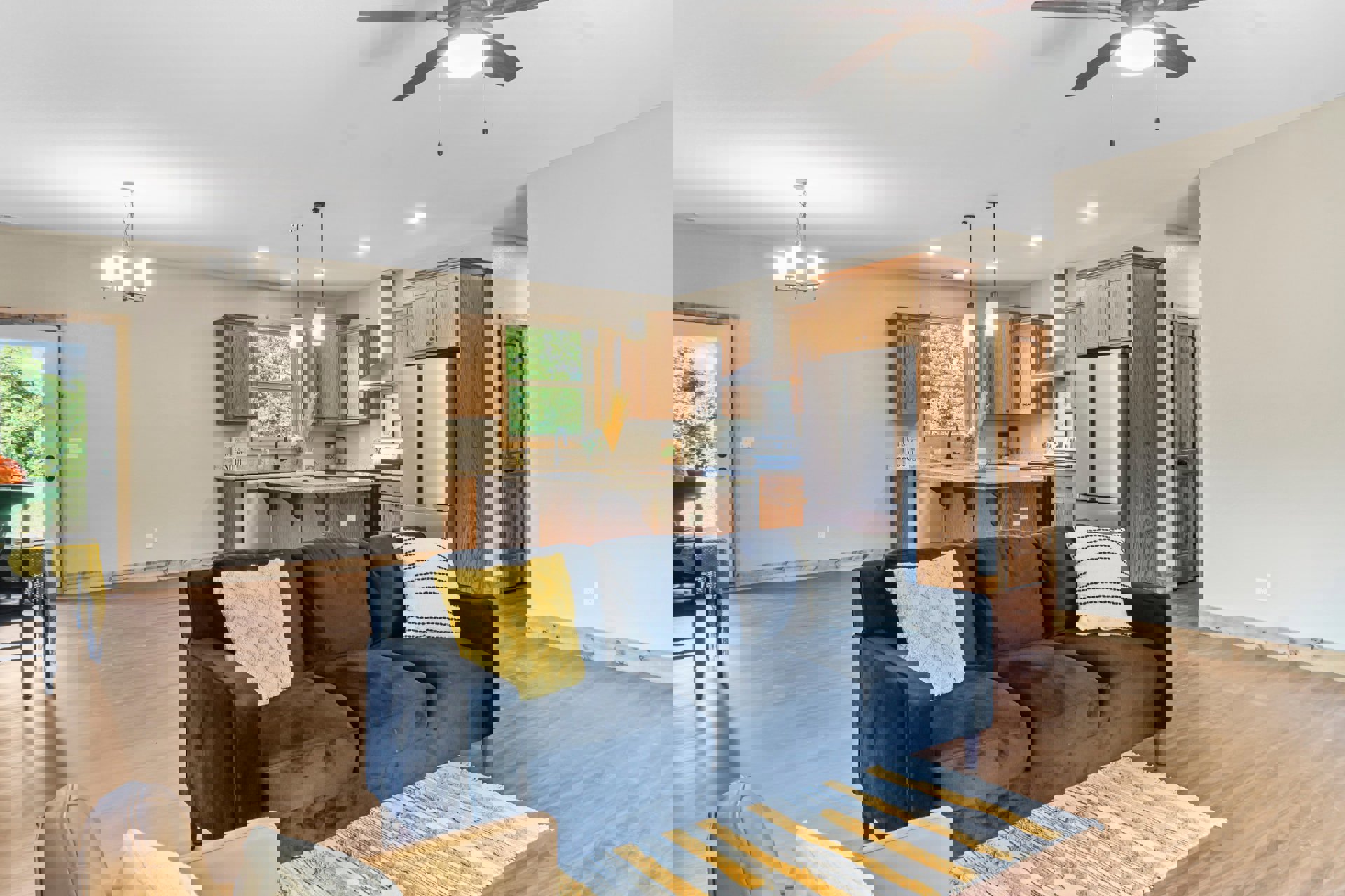 Open-concept living room with light gray couch, patterned and yellow accent pillows, blue chair, potted plant, and modern kitchen featuring white cabinetry and refrigerator with