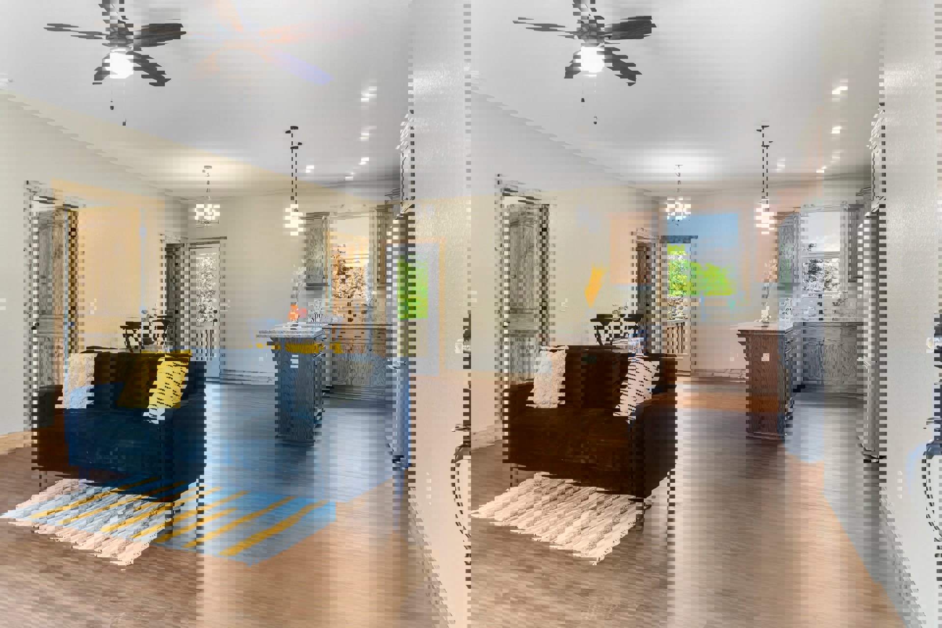 Open-concept living room with gray couch, assorted pillows, black accent chair with yellow pillow, patterned area rug, laminate flooring, and adjacent kitchen; ceiling fan with