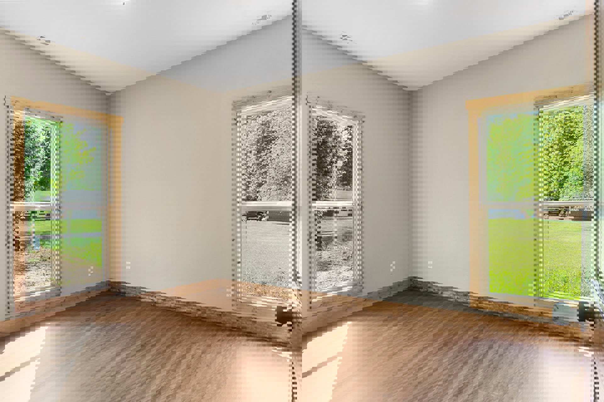 Spacious room with light wood flooring, large windows overlooking green yard and trees, white walls, natural daylight filling the space