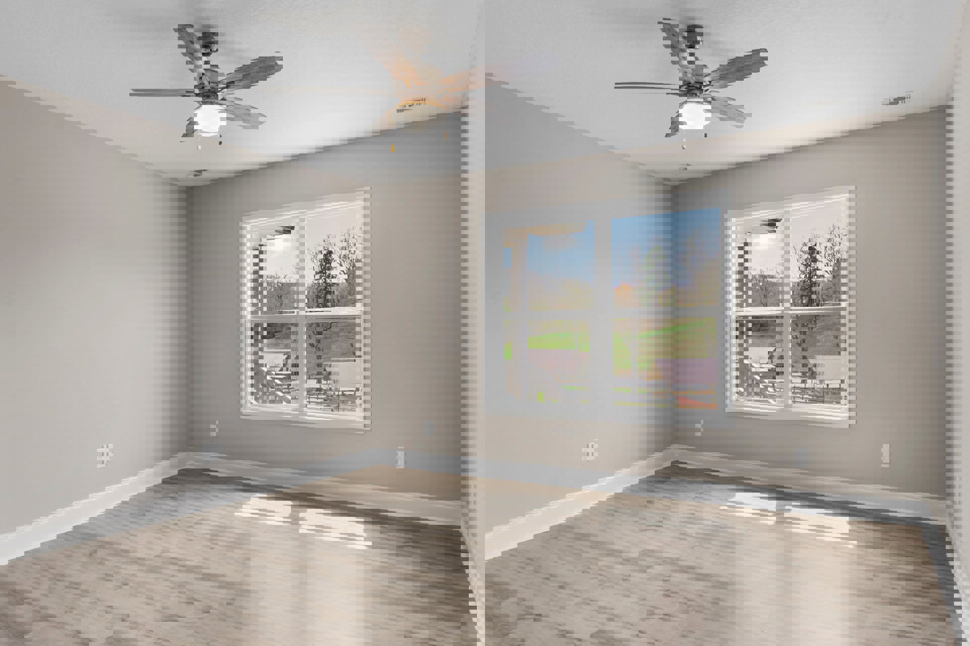 Ceiling fan with integrated light fixture mounted on white plaster ceiling, large window overlooking landscape, sunlight streaming onto wood flooring in residential interior.