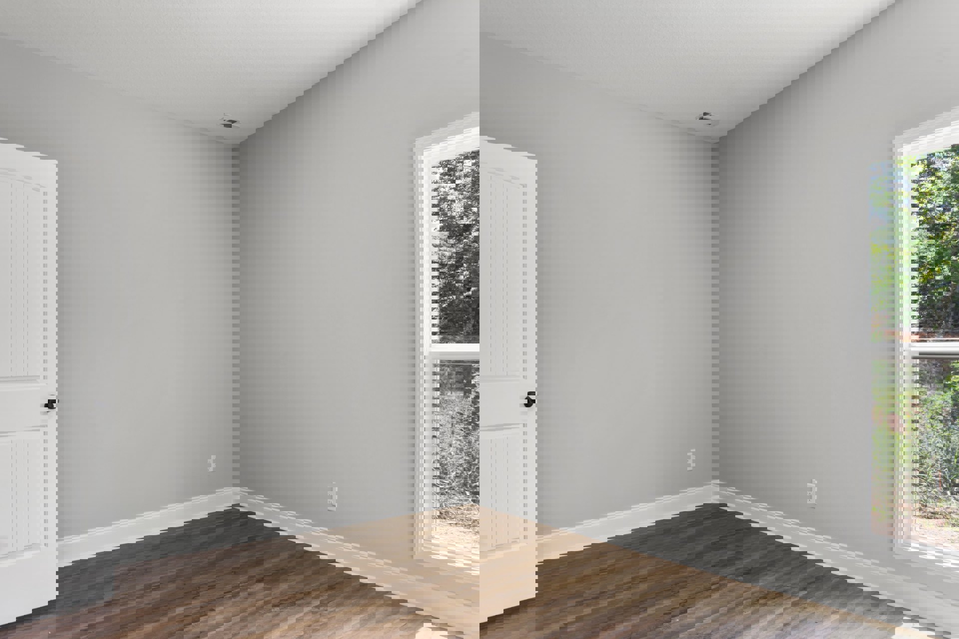 Wood floor with white trim, white walls, white door with black knob, window overlooking trees, white ceiling with air vent