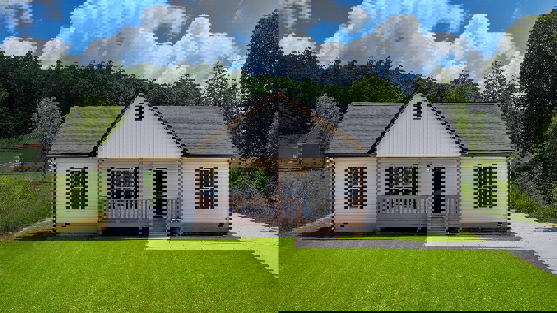 White siding house with covered porch, black front door, wooden railing, green grass lawn, concrete walkway, and multiple windows.