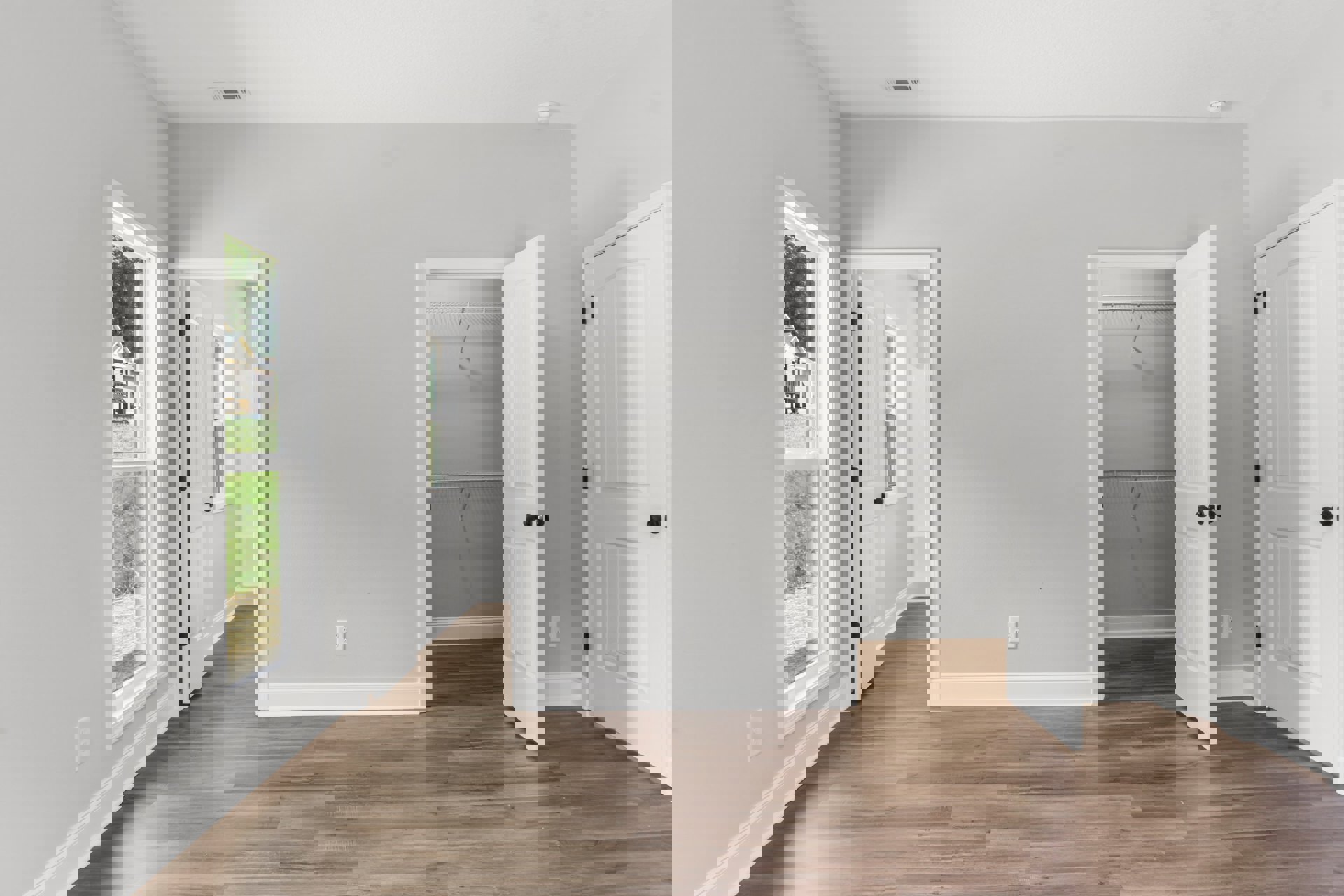 Open white doors with black knobs leading to a room featuring wood flooring, a white closet with shelving, a window overlooking a deck and grassy yard, and a light switch on the