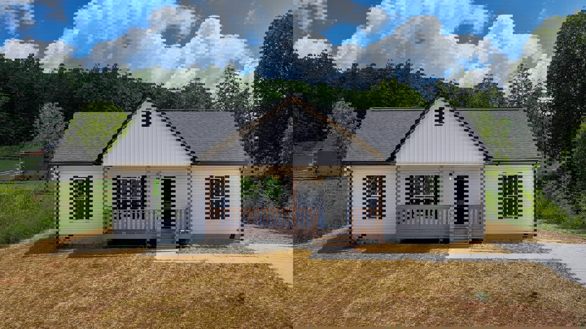 White house with covered porch, white railing, grassy lawn, and small cottage in the background; trees and cloudy sky visible.