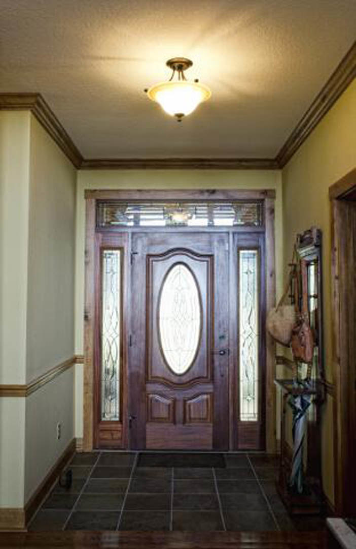 White paneled hallway door with brushed metal handle, overhead ceiling light fixture, beige tile flooring, partial view of a person’s leg, and white wall molding