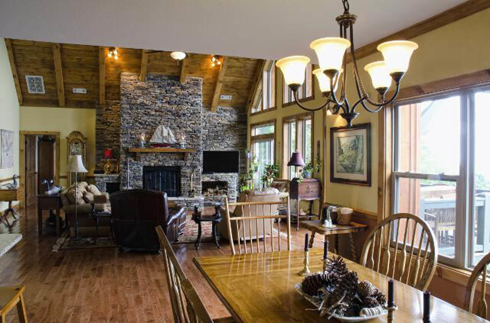 Living room with stone fireplace, wooden table topped with pine cones and candles, leather chair in foreground, framed artwork on wall, chandelier overhead, hardwood floors