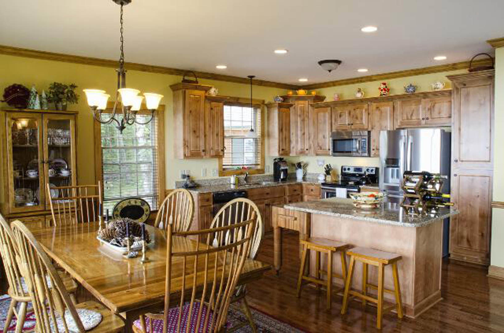 Open kitchen with wood cabinetry, stone countertops, dining table set with pine cones and candles, wooden chairs, and kitchen island with stools and bowl of food; large window in