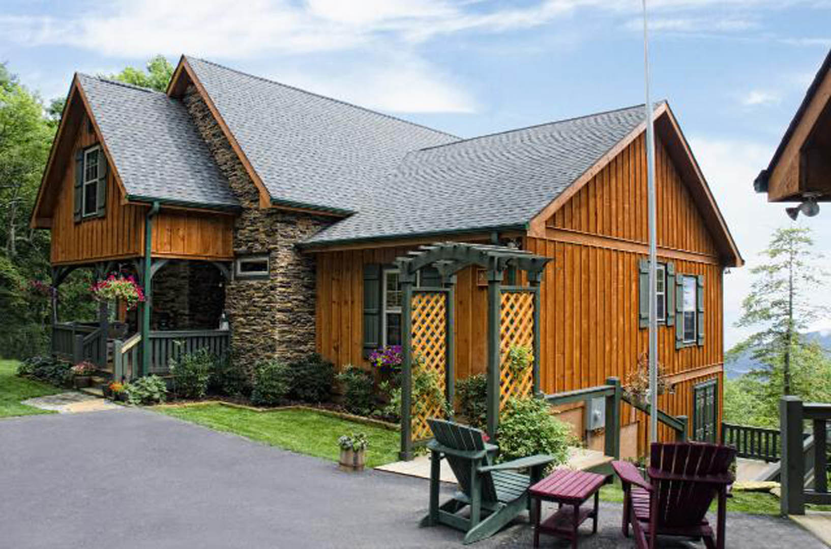 Stone exterior walls, green chair and red table on patio, wooden fence with trellis, cloudy sky, plants surrounding home