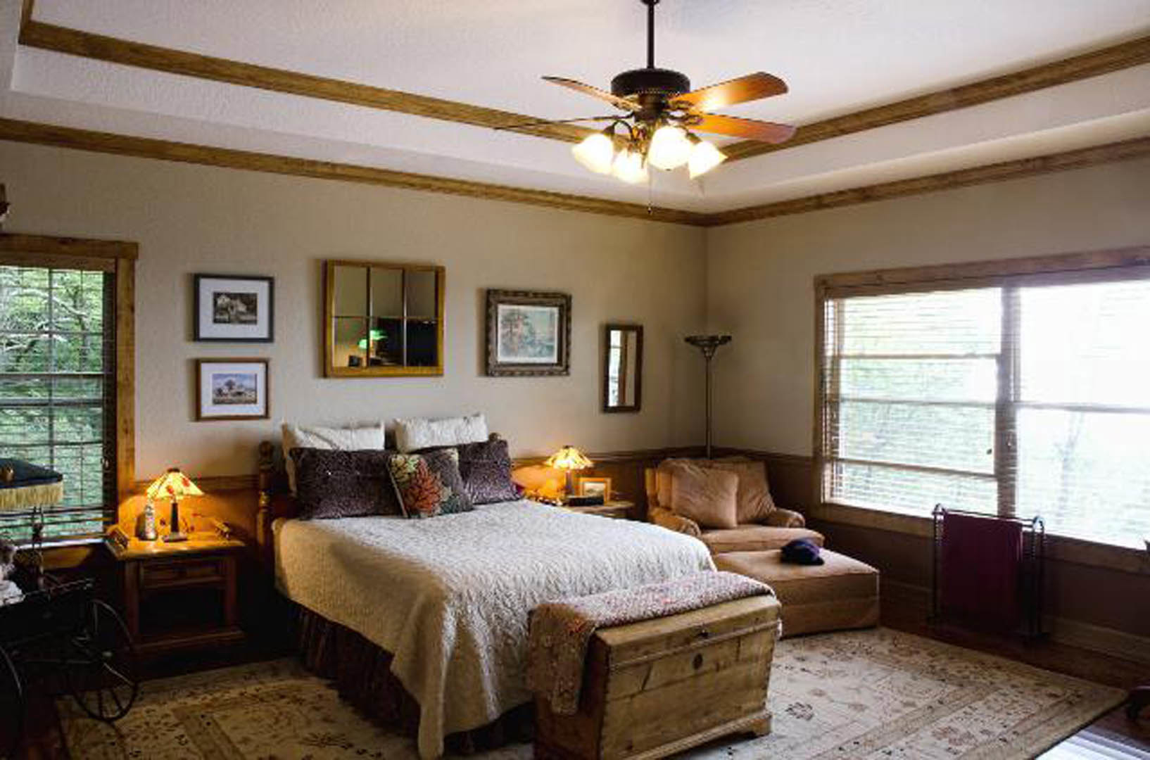 Bedroom with a ceiling fan, upholstered bed with pillows, bedside lamp, wooden chest topped with a blanket, couch with accent pillows, towel hanging on a rack, window reflecting
