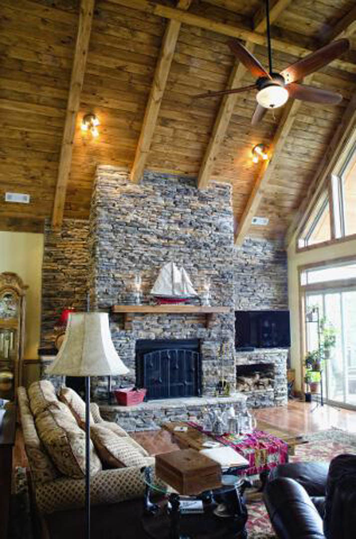 Living room with stone fireplace, exposed wooden ceiling beams, neutral couch with accent pillows, leather chair, hardwood floors, and soft lighting
