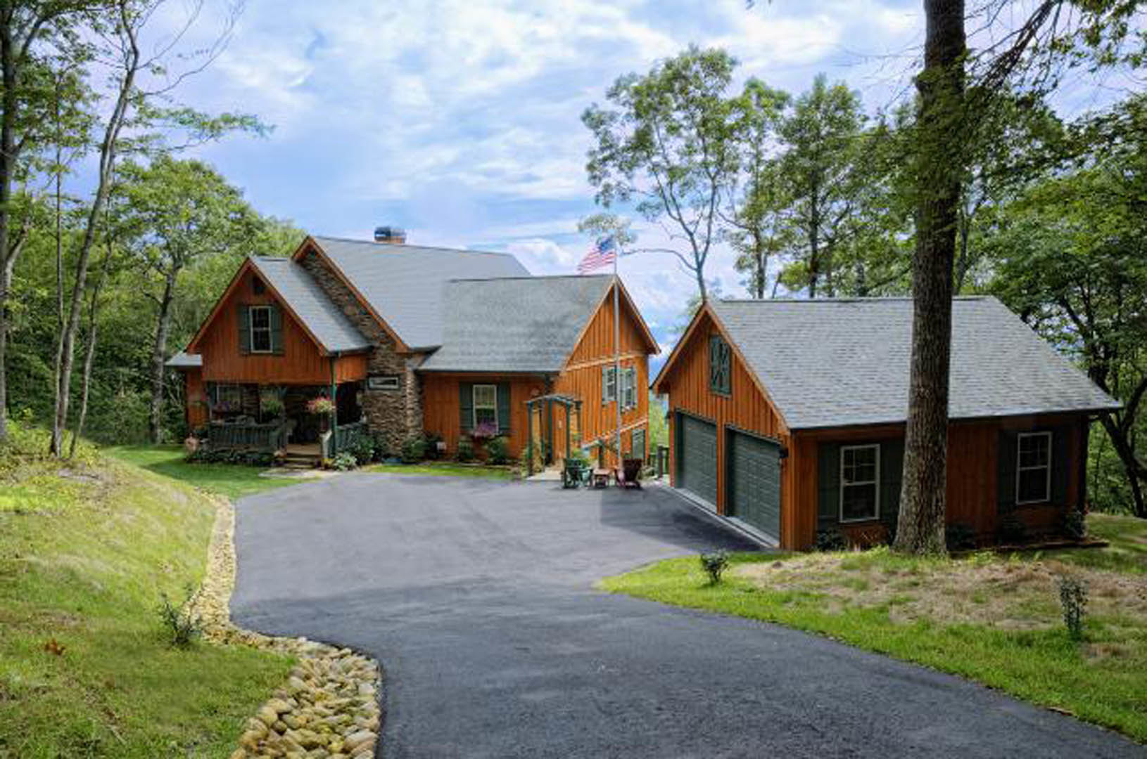 Two-story home with stone porch and stone wall, concrete driveway bordered by green lawn, mature trees in background, cloudy sky overhead