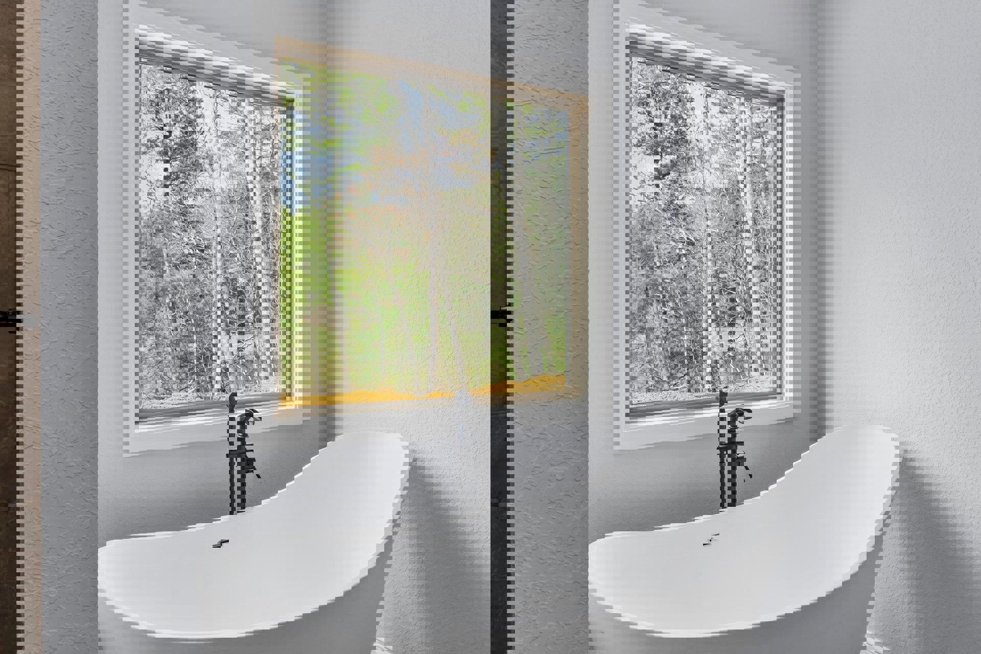 Freestanding white bathtub with matte black faucet beneath large window overlooking trees, surrounded by light-colored walls in modern bathroom