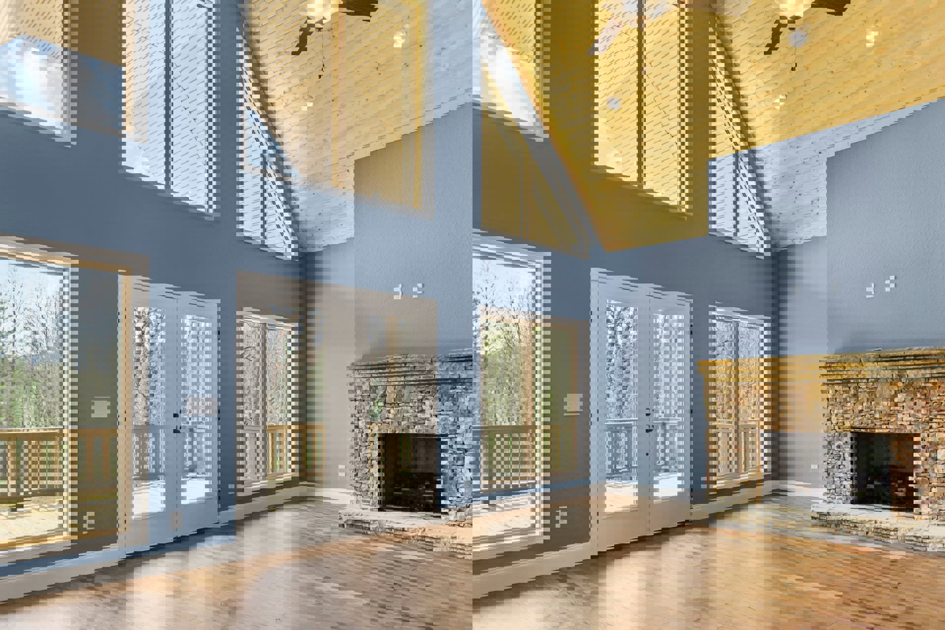 Hardwood floor living room with stone fireplace, blue accent wall, ceiling fan, glass-paneled double doors, and window overlooking trees and balcony