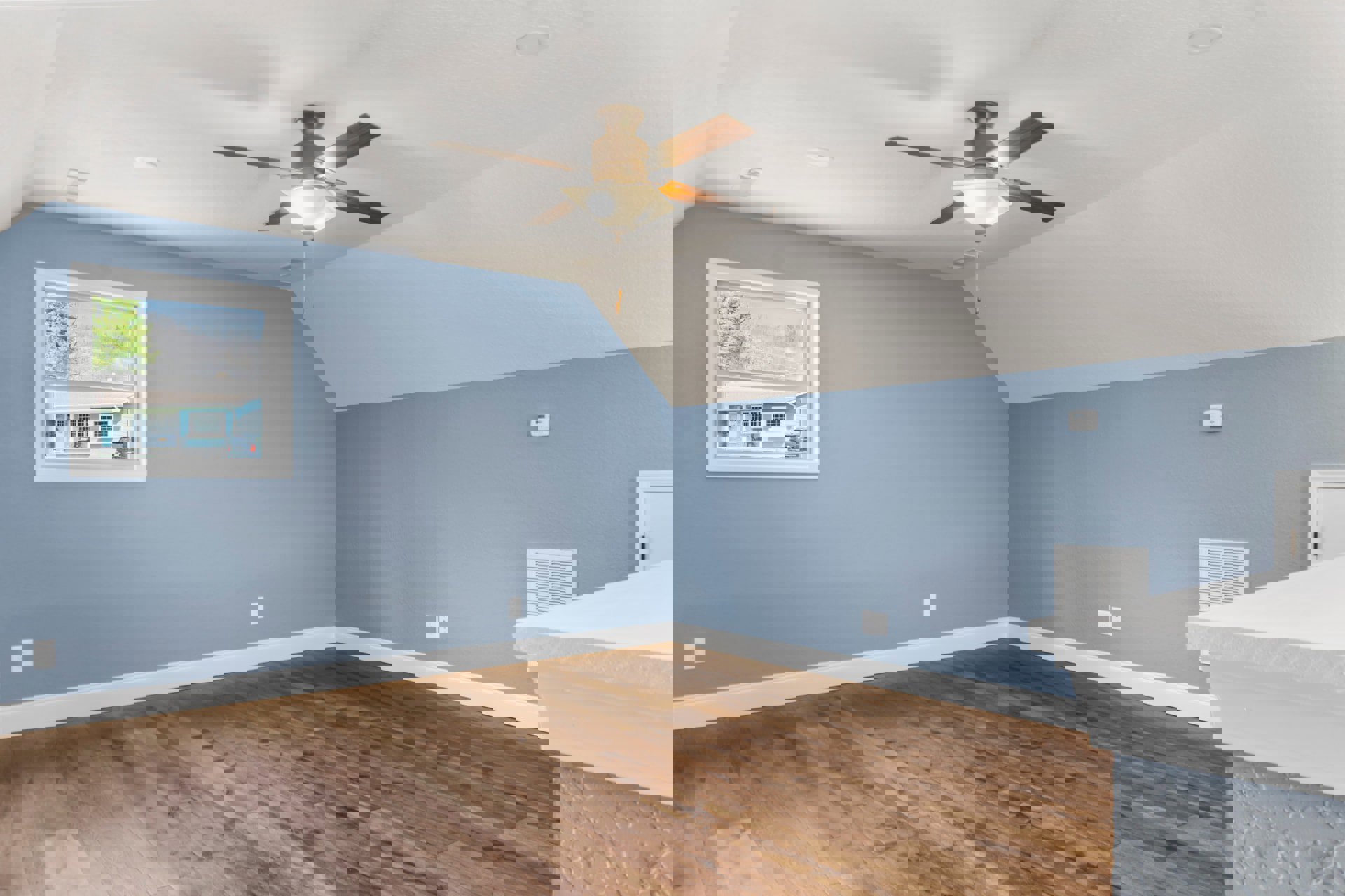 Living room with blue walls, patterned hardwood floor, ceiling fan with light, white fireplace, and large window overlooking blue house and trees.