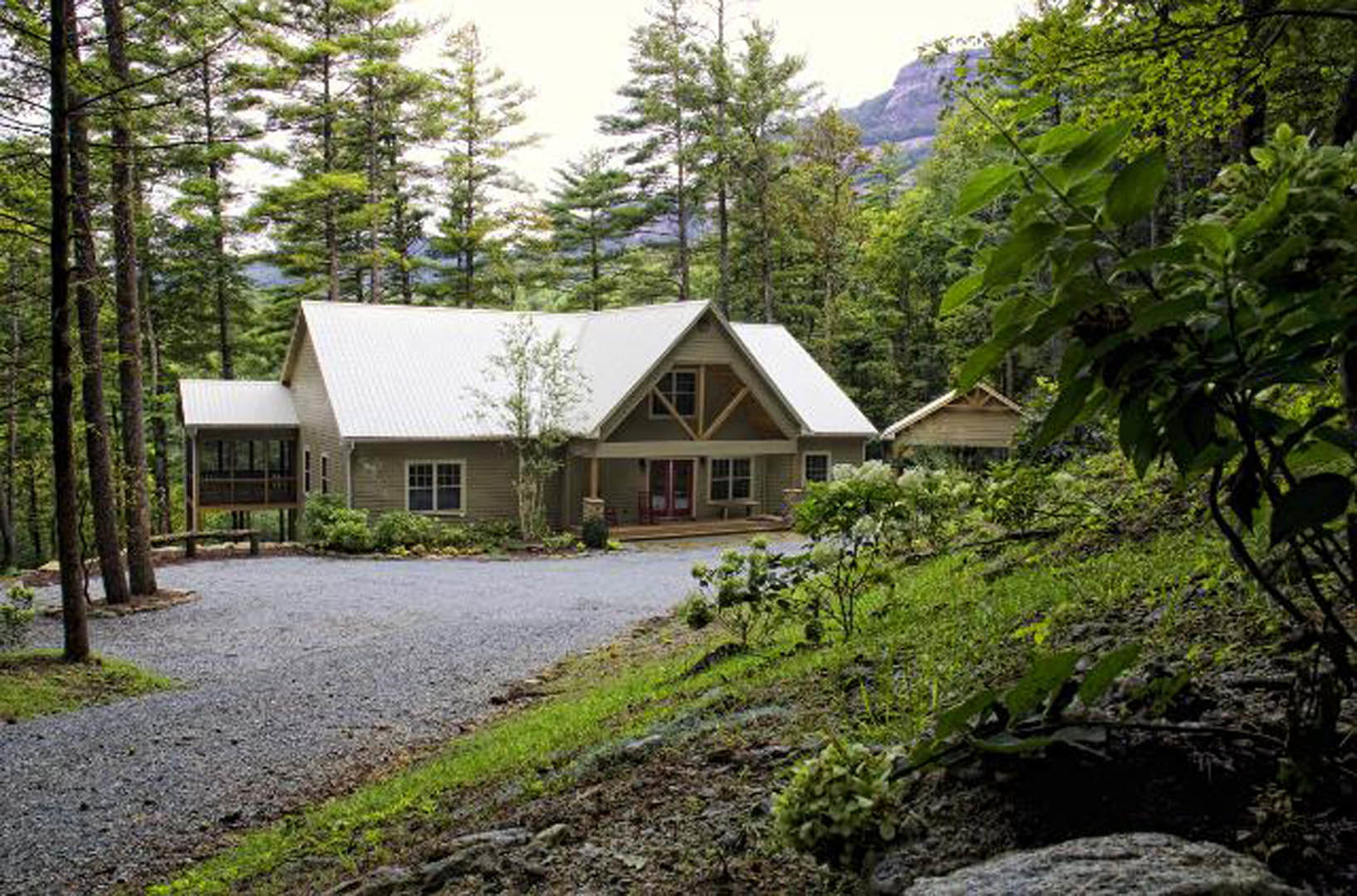 Wooded setting with a gravel driveway leading to a farmhouse-style home, white-framed windows, pitched roof, and mature trees surrounding the exterior