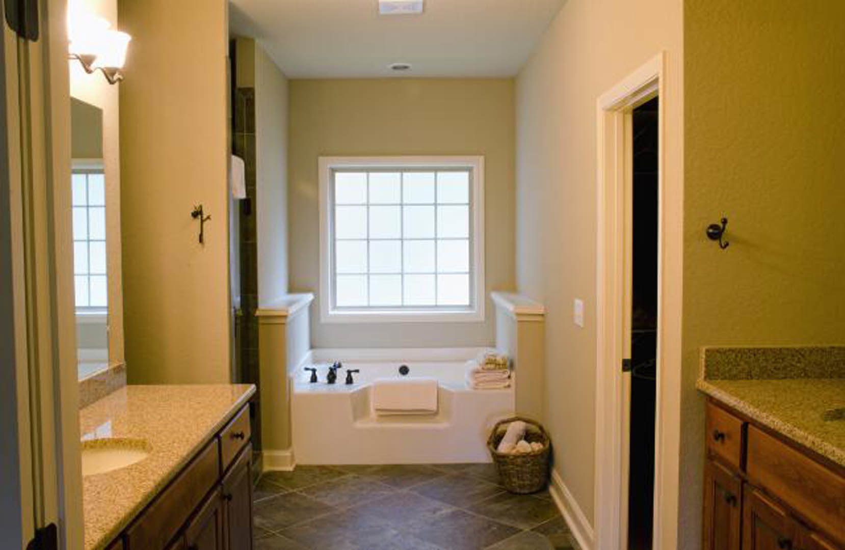 Freestanding white bathtub beside a multi-pane window, white sink with black faucet on yellow vanity, black wall hook, basket filled with rocks, grey tile flooring