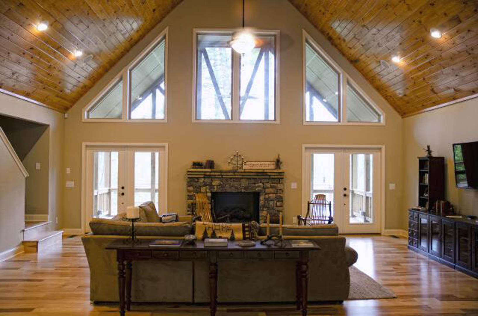 Living room with hardwood floors, stone fireplace, gray couch, wooden coffee table with a book, large window with sunlight, white ceiling with recessed lighting, built-in