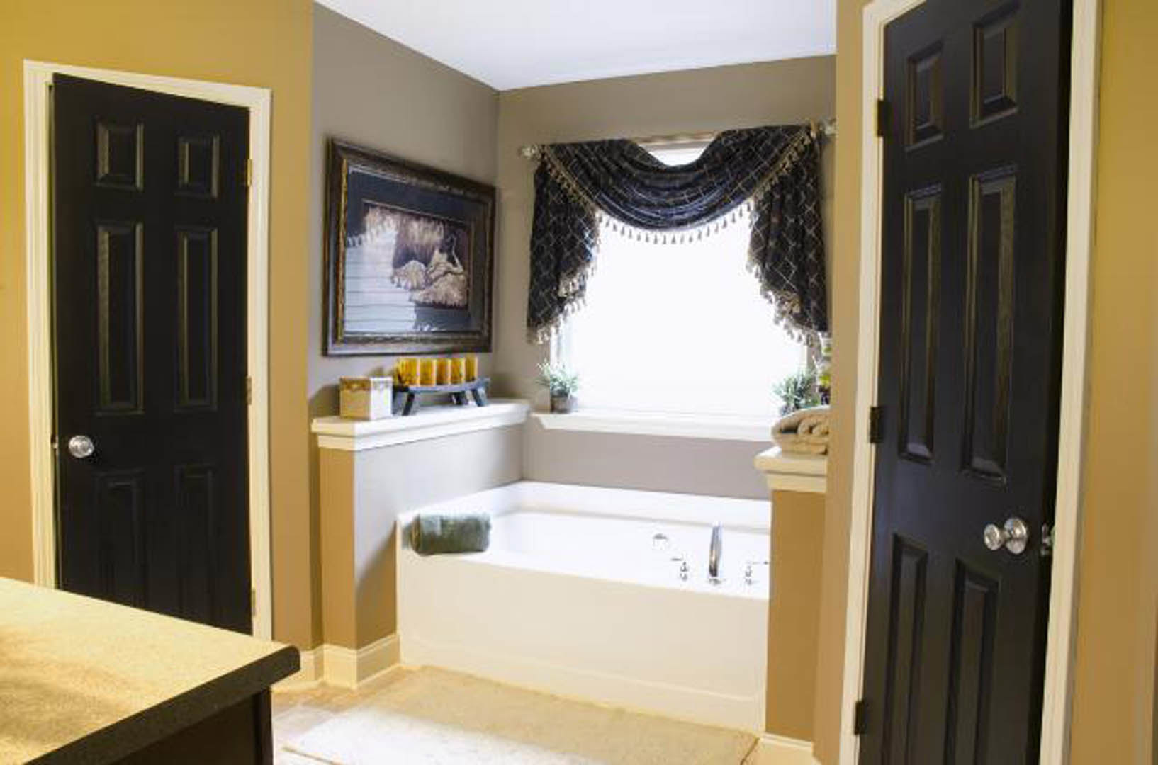 White freestanding bathtub beneath a window with sheer curtains, dark cabinetry, wall-mounted mirror, framed cat artwork, and black door in a modern bathroom