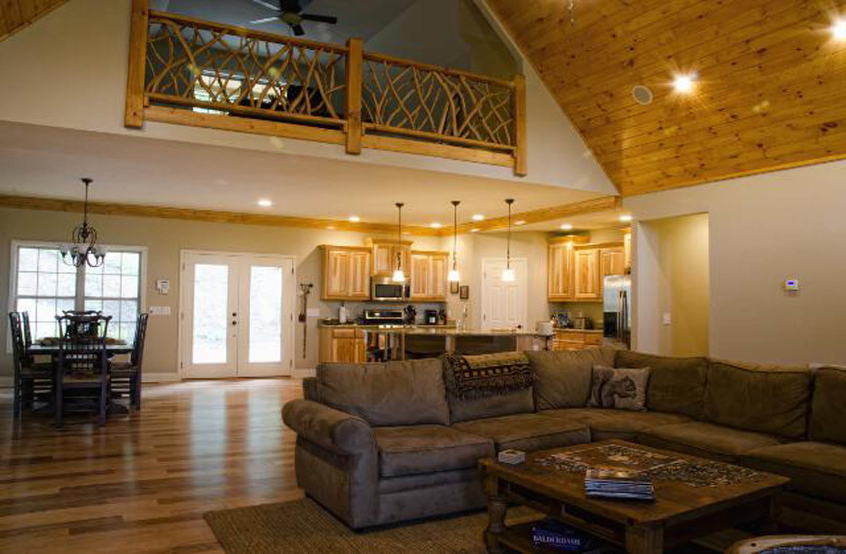 Open concept living room with light-colored couch, wooden coffee table, and adjacent kitchen featuring glass double doors, balcony with wooden railing, and dining table with