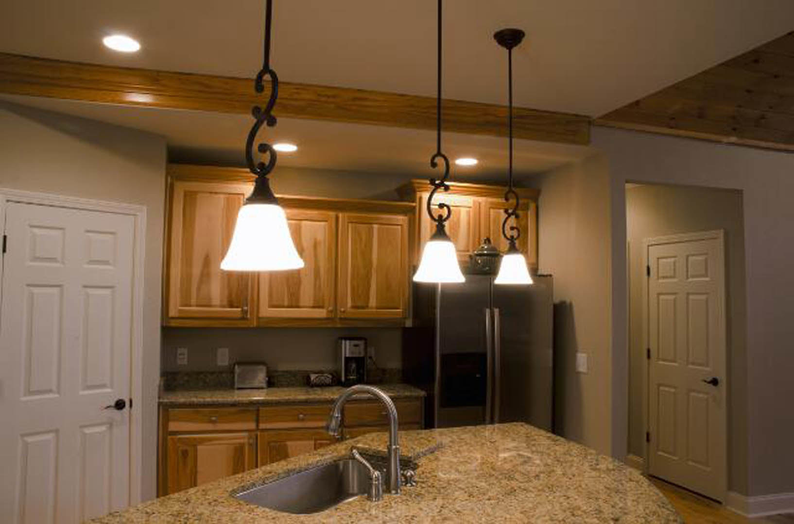 Modern kitchen featuring white shaker cabinets, stainless steel sink with chrome faucet, light-colored stone countertops, two pendant lamps above a stainless refrigerator, and a