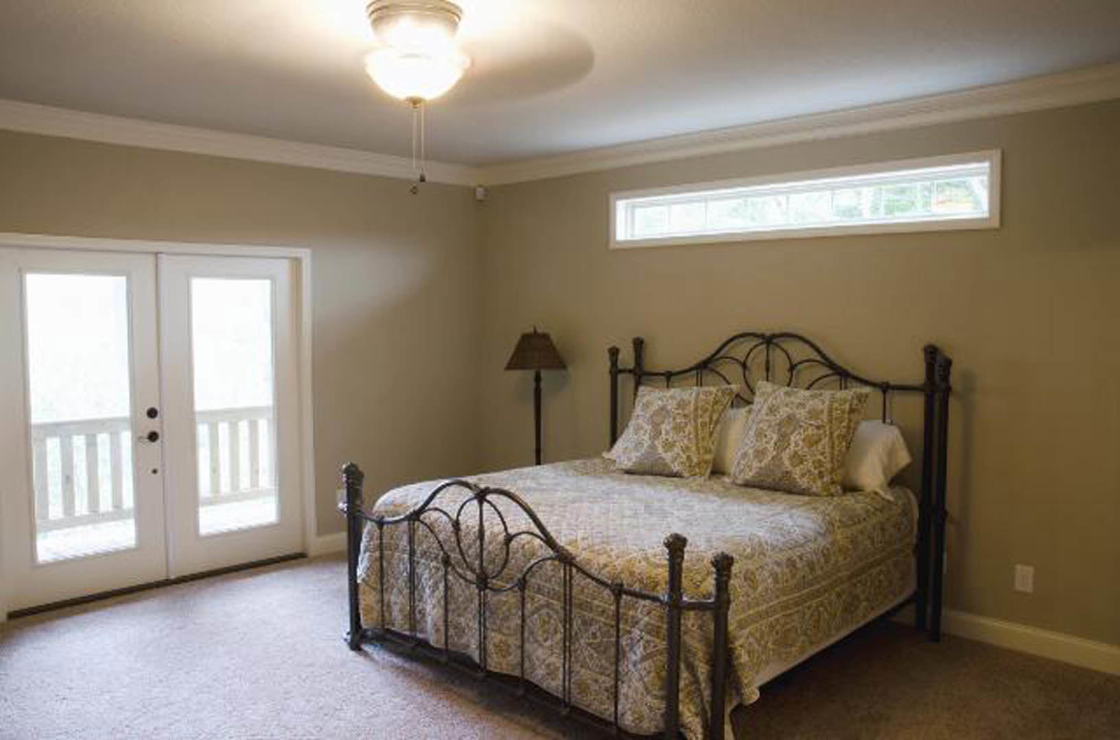 Metal bed frame with white bedding, square-shaded lamp on bedside table, double glass-panel doors, window with soft light, ceiling light fixture, hardwood floor