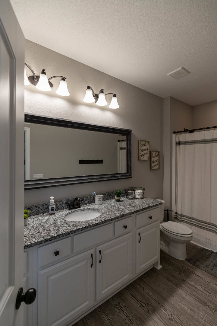 Bathroom with white tile walls, rectangular mirror above a white sink and countertop, row of wall-mounted lights, white toilet, white shower curtain, and black-framed board.