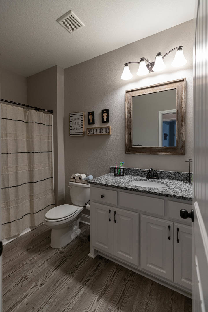 Bathroom with white tile walls, wooden-framed mirror above a stone countertop sink, modern faucet, three-light fixture, toilet, neutral cabinetry, and a decorative wall sign.
