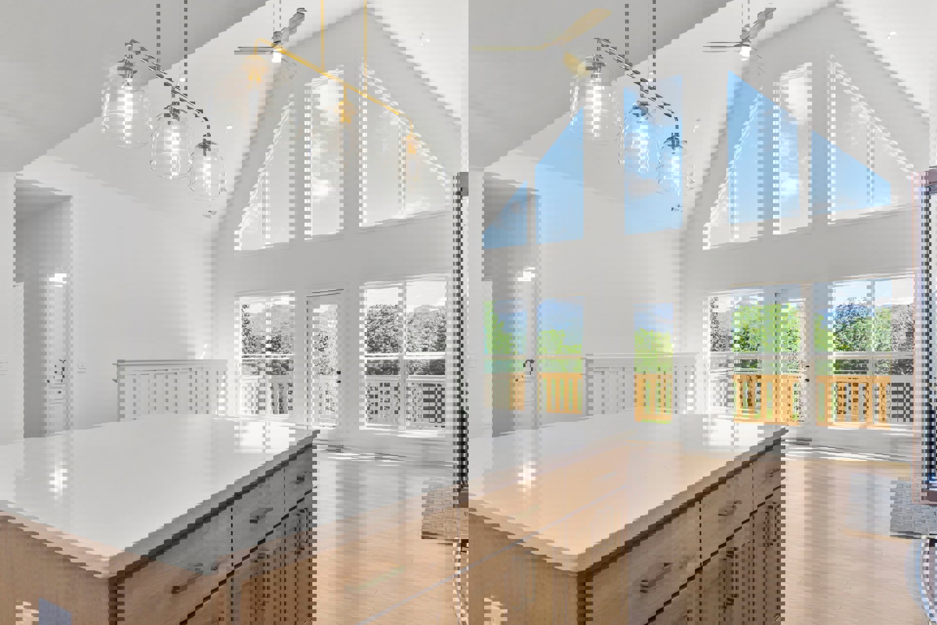 Spacious kitchen featuring a large white island with wooden drawers, stainless steel sink and tap, ceiling fan with light, clear glass pendant fixture, window framing blue sky and