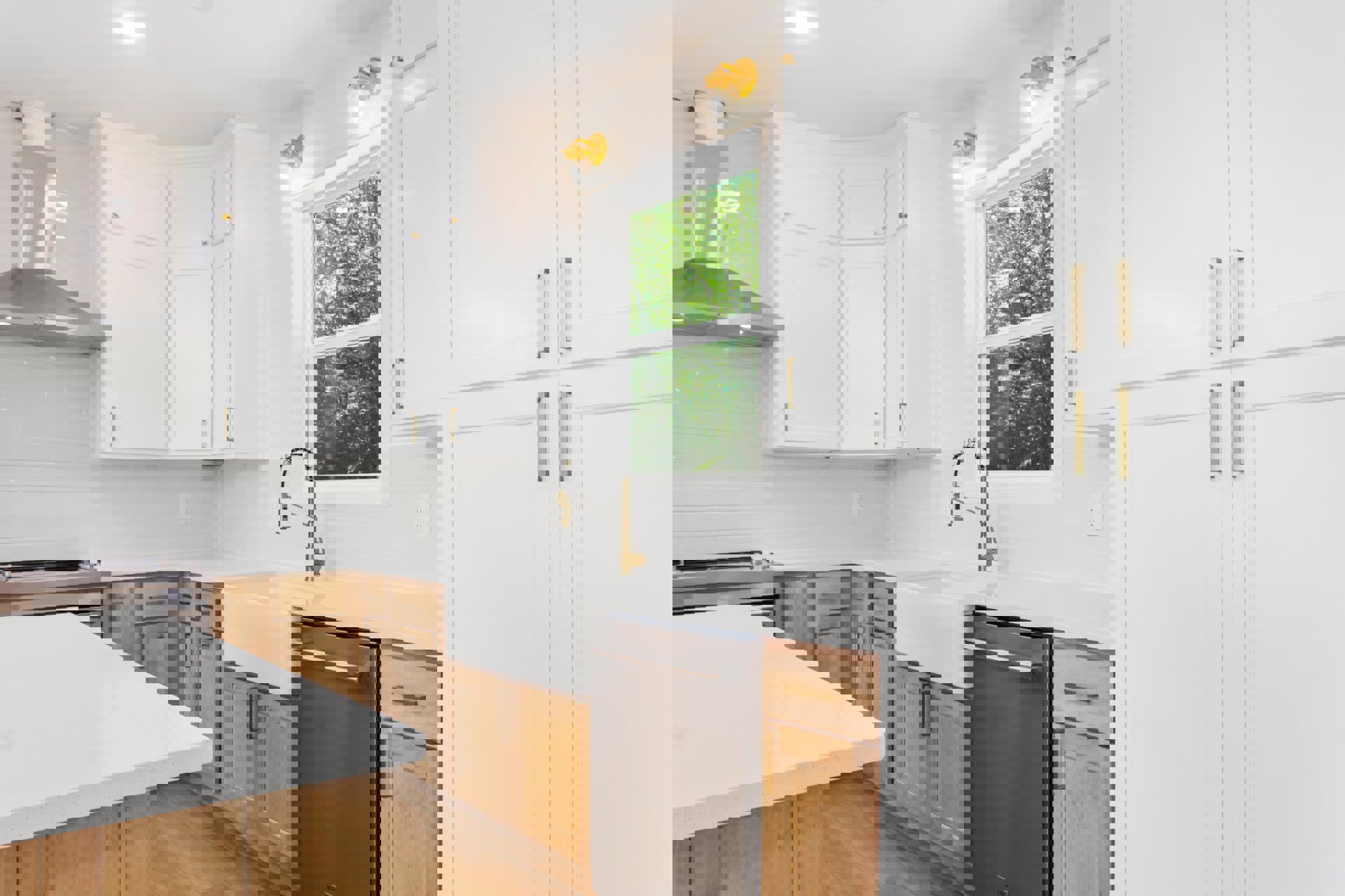 White kitchen cabinets, gold and black faucet, white countertop with sink, built-in dishwasher, wall-mounted light fixture, window overlooking trees.