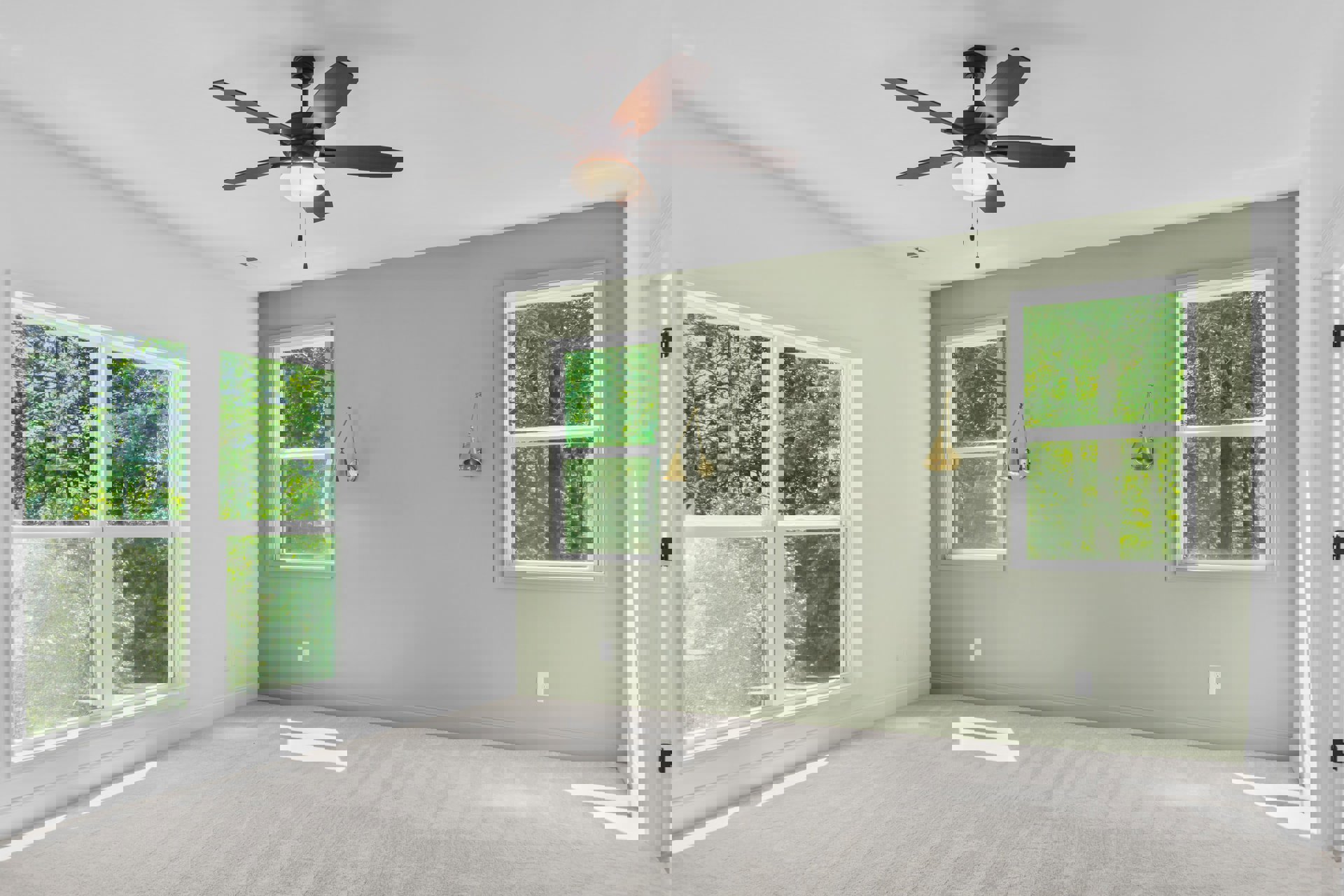 Ceiling fan with light fixture above white carpeted floor, two large windows with white frames showing green trees outside, neutral walls and close-up of lamp in corner