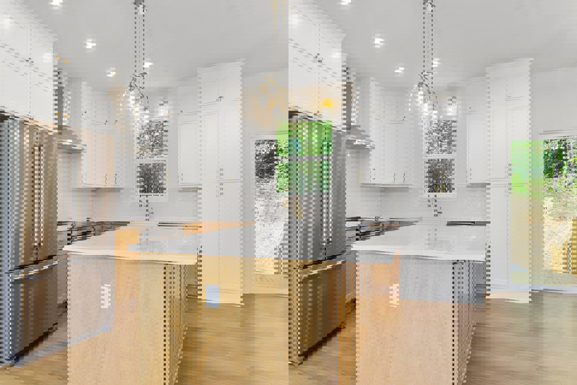 Spacious kitchen featuring a large island with a white countertop, white cabinetry with gold handles, stainless steel refrigerator, wood surface with a white outlet, and modern