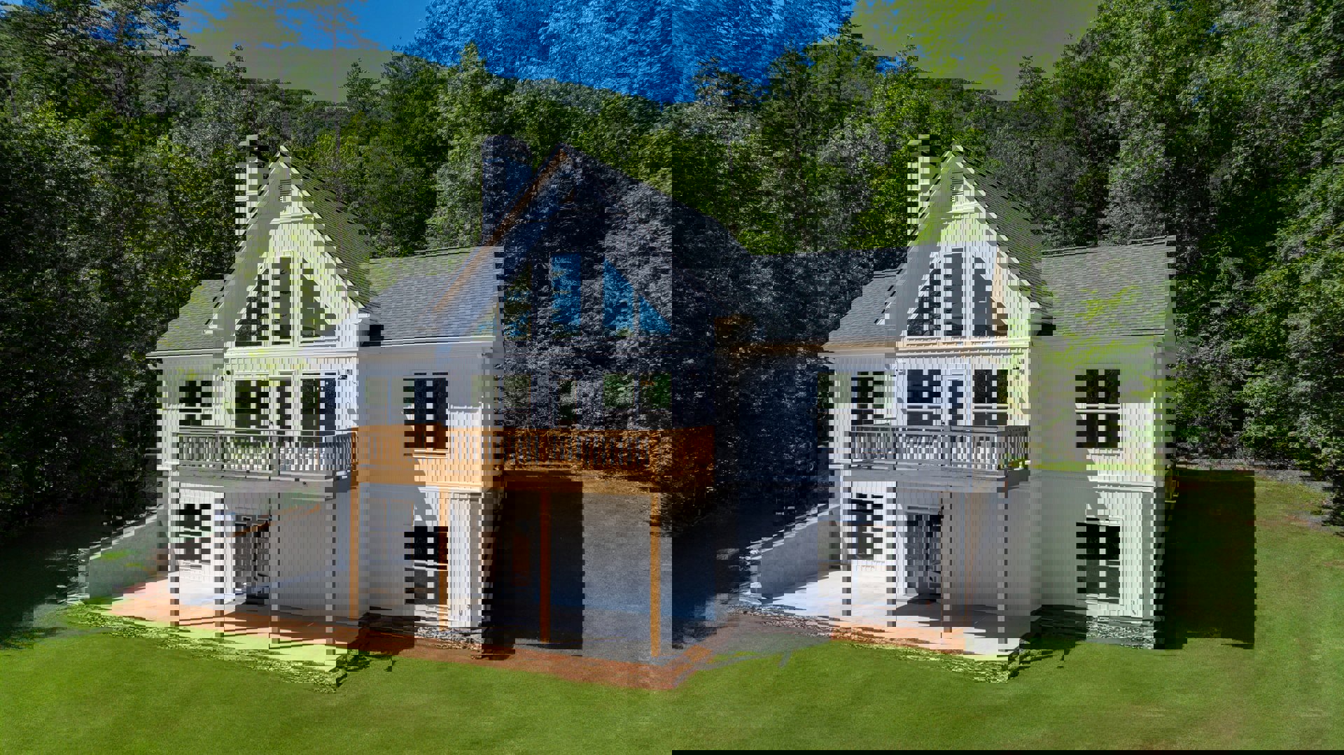 White two-story house with white-framed windows, second-floor balcony with metal railing, manicured green lawn, leafy tree branches, and clear blue sky