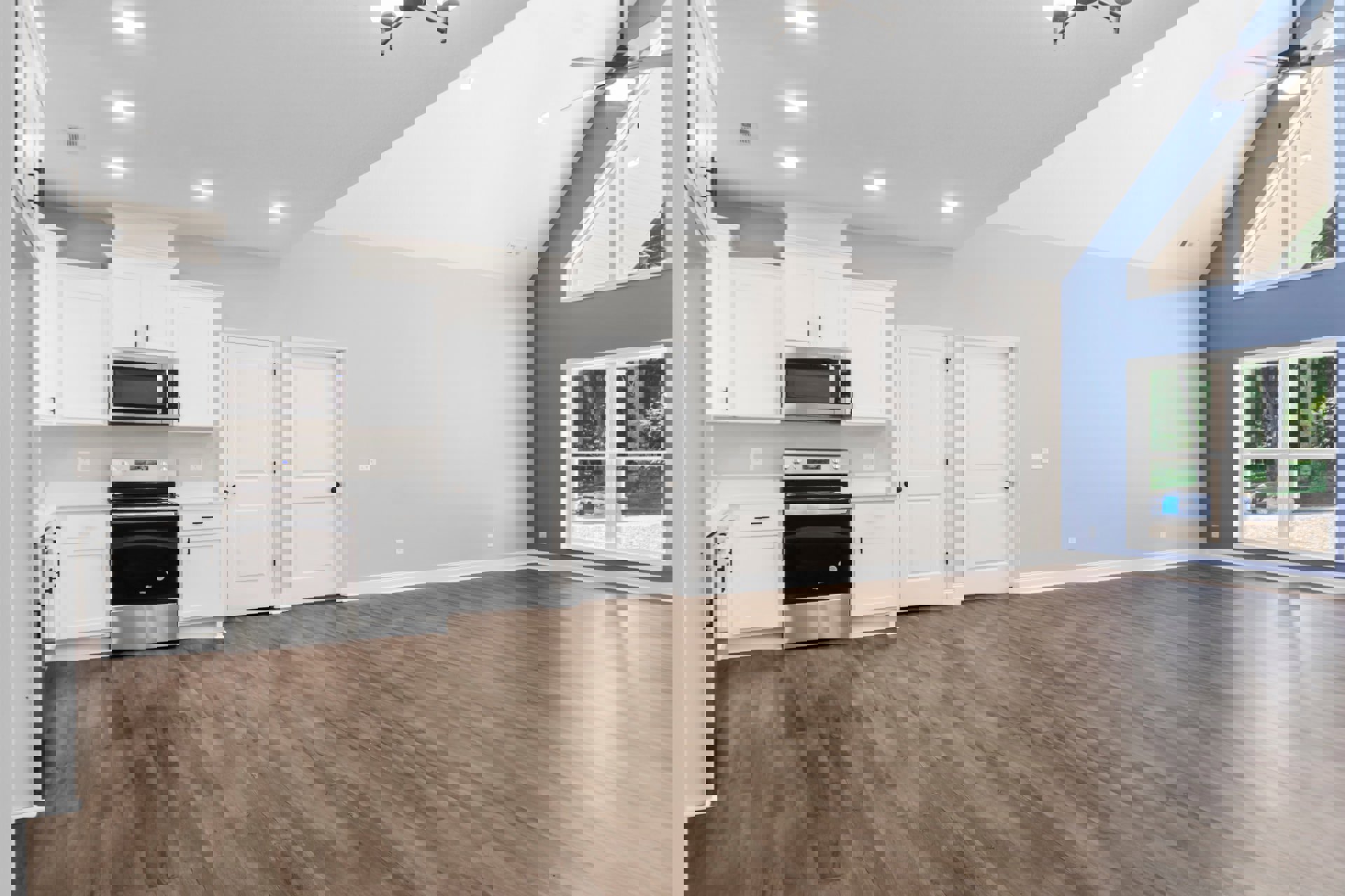 Open kitchen and living room featuring wood floors, white cabinetry, stainless steel stove and microwave, white doors with black handles, large window with blue bucket visible