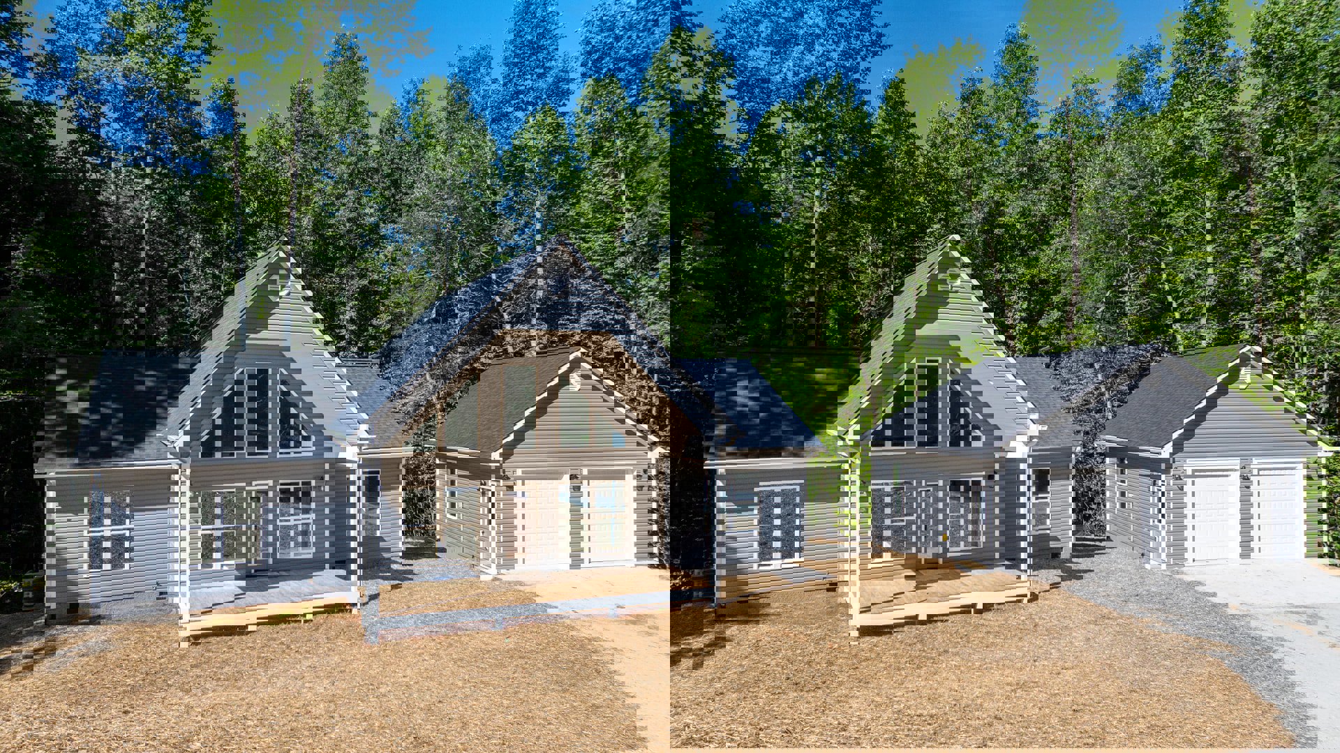 Two-story home with white trim, covered front porch with wood decking, gray garage doors, concrete driveway, and landscaped yard bordered by mature trees.