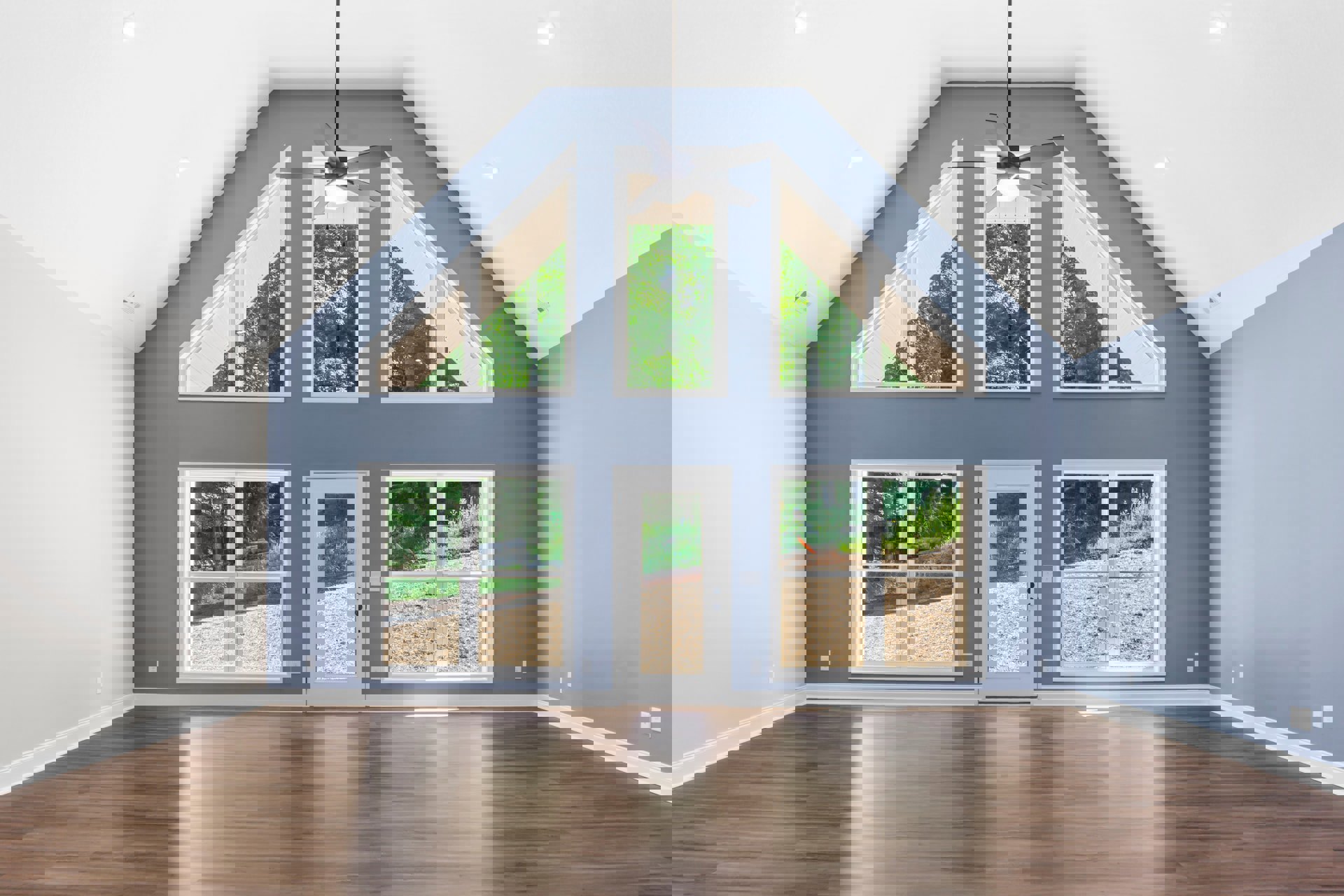 Living room with hardwood floors, ceiling fan with light, open door featuring glass window, large windows overlooking garden and forest.