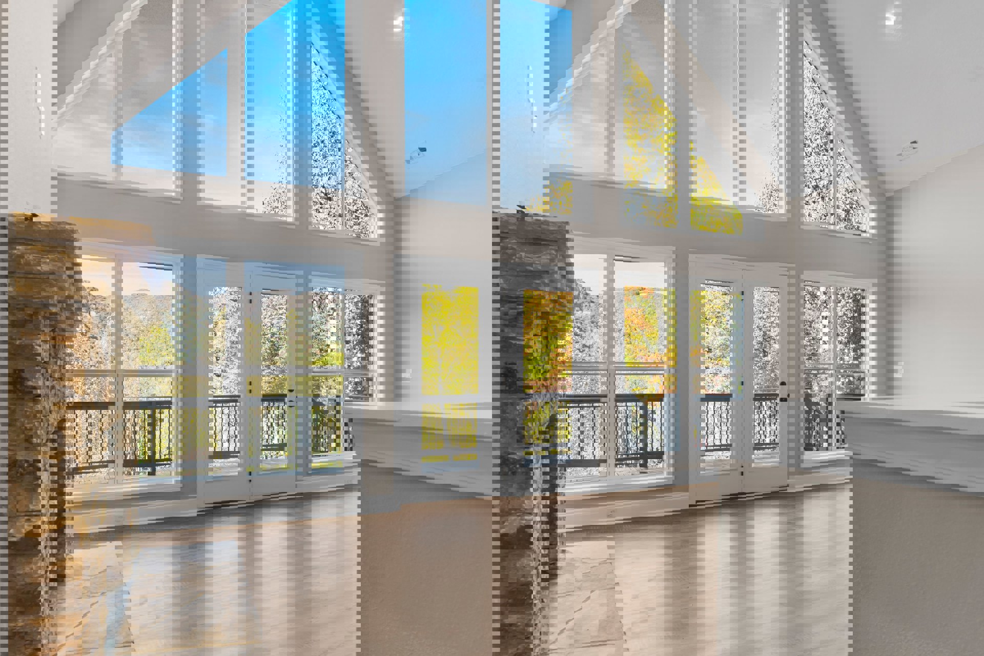 Spacious interior with wide plank wood flooring, stone fireplace beside a window, large white kitchen island, double glass doors opening to deck, tree with yellow leaves visible