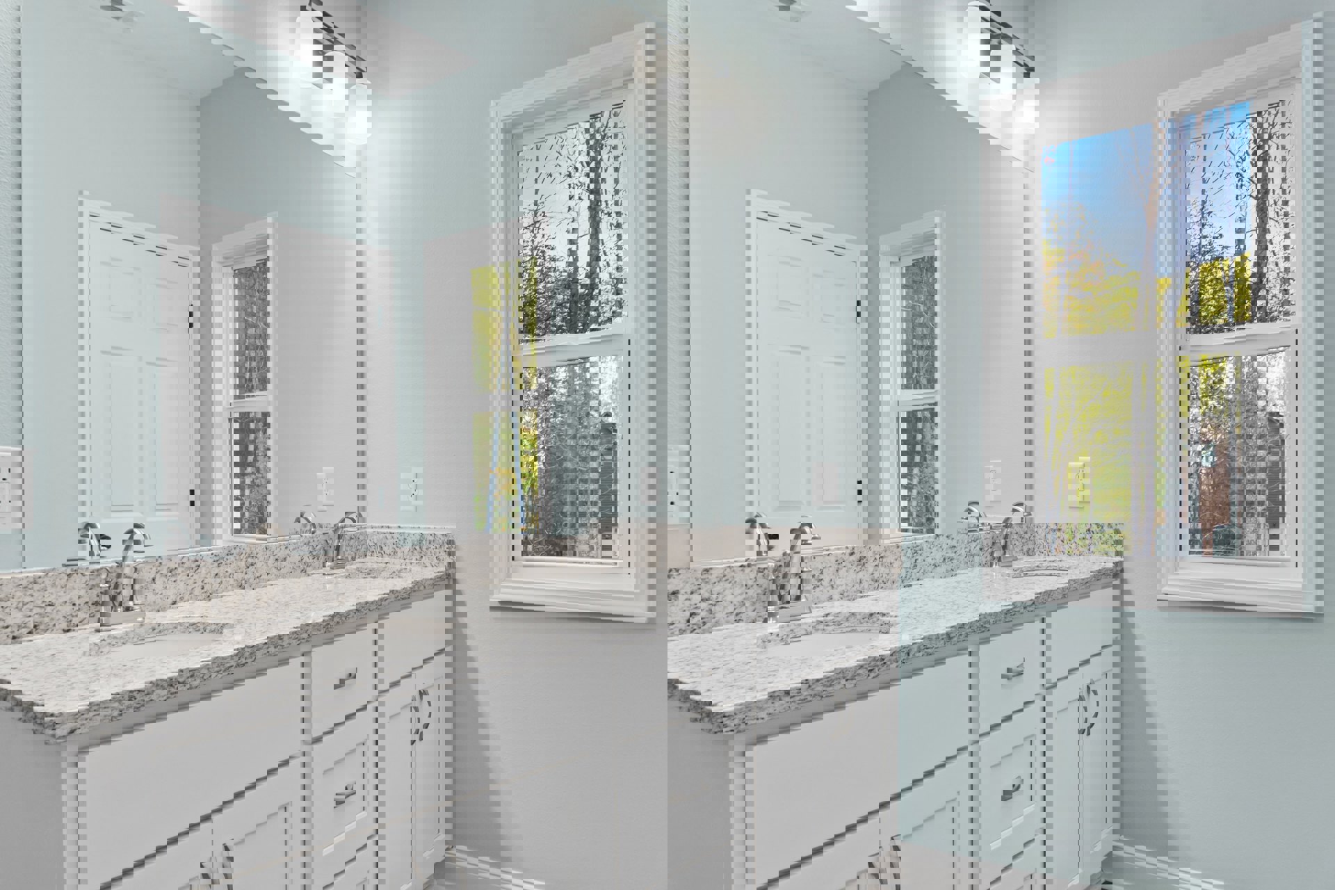 White bathroom with marble countertops, white cabinets, chrome faucet, white door featuring a cross detail, window overlooking trees, modern light fixture above sink.