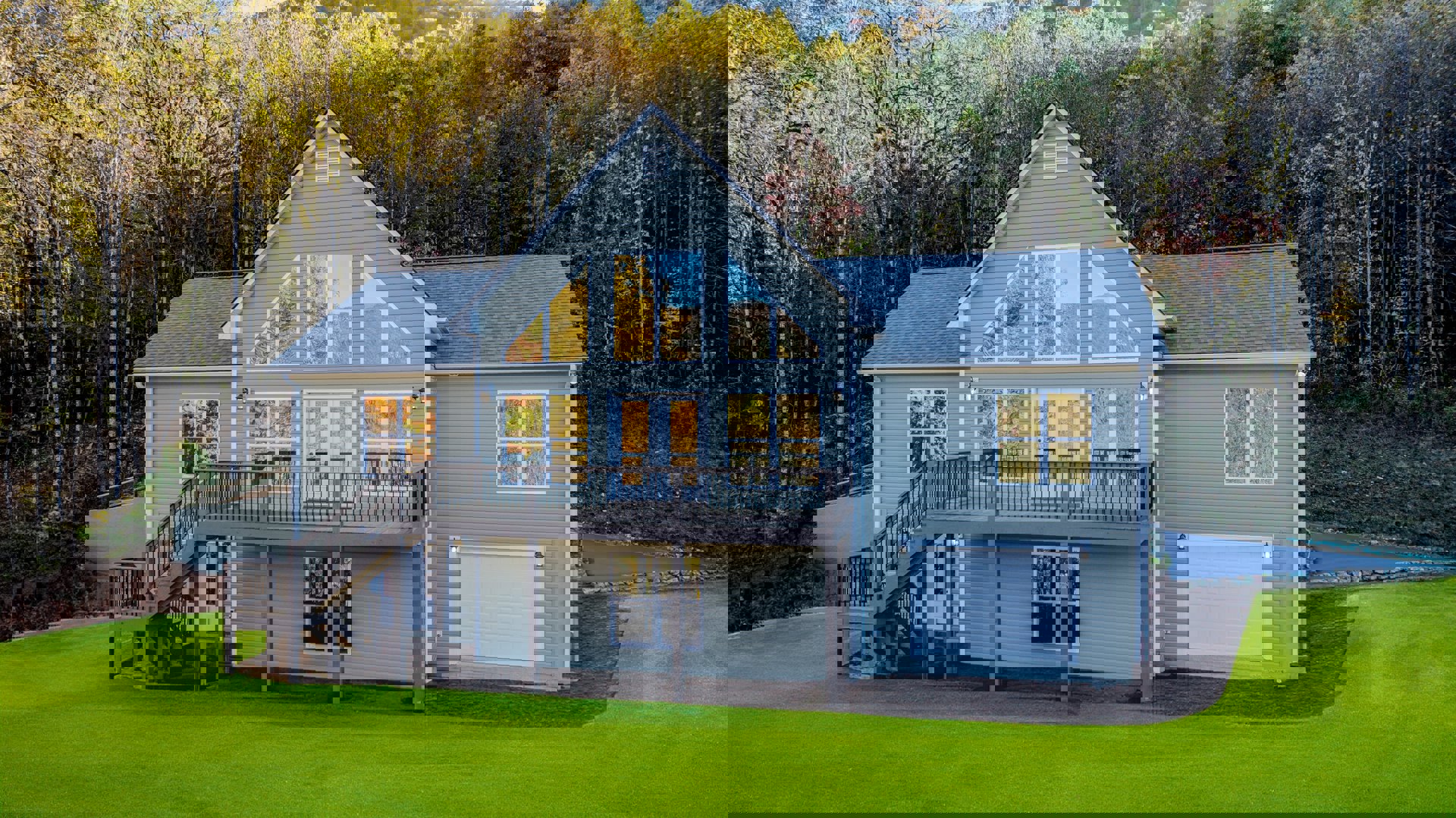 Two-story house with white siding, attached garage, front porch, wooden deck, green lawn, and mature trees in the background