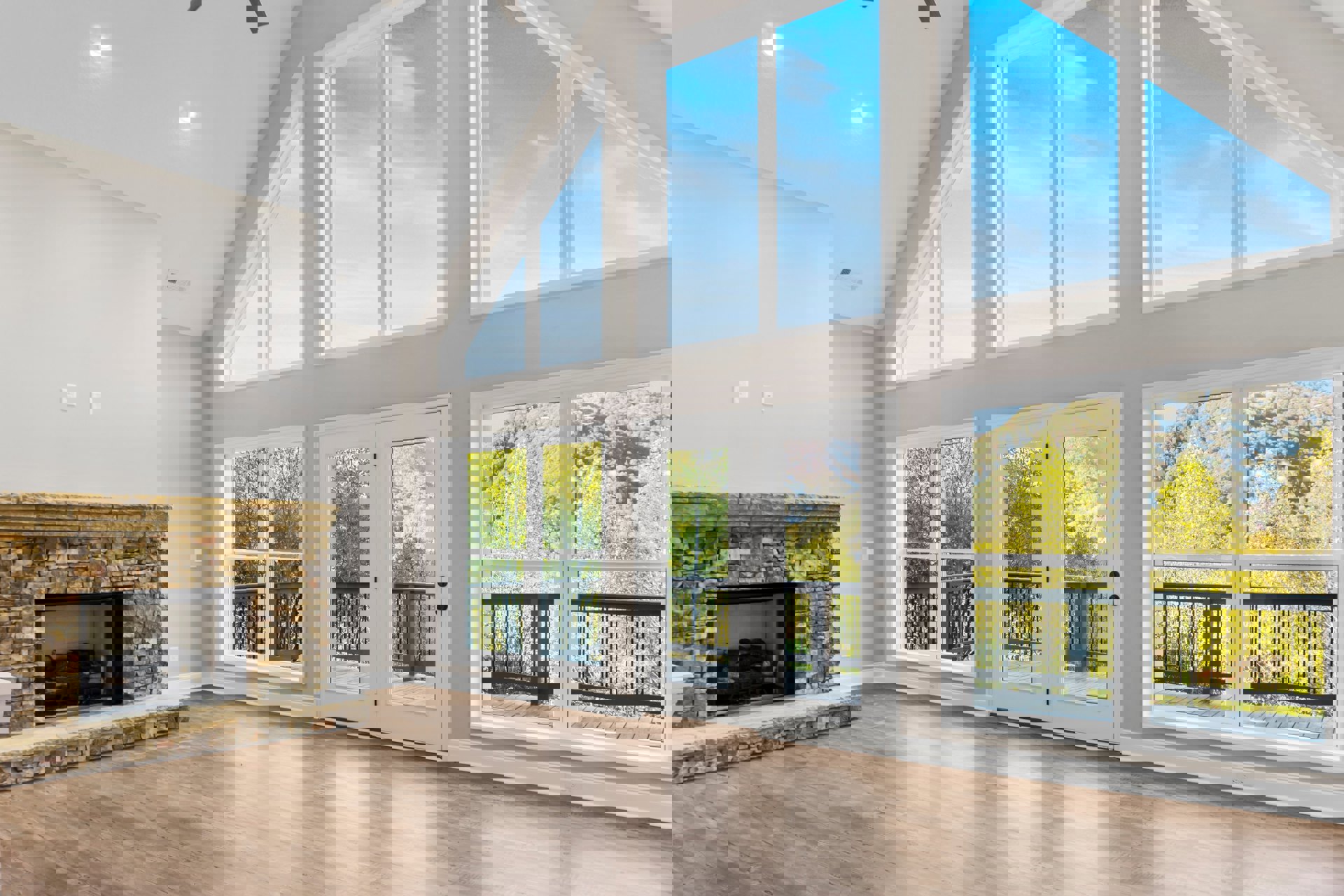 Living room featuring a stone fireplace with stacked logs, hardwood floors, large windows overlooking a deck, and a white door with glass pane.