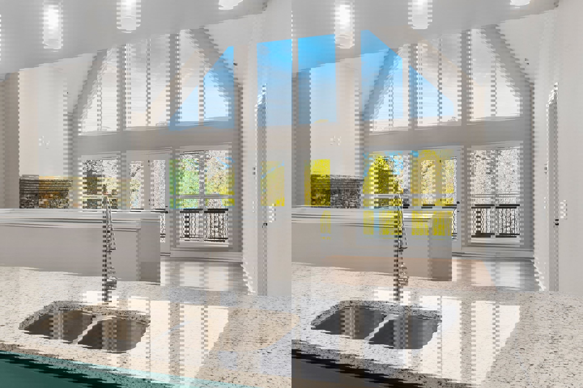 Granite countertop with double stainless steel sink and modern faucet beneath a large window, stone tile backsplash, and neutral cabinetry