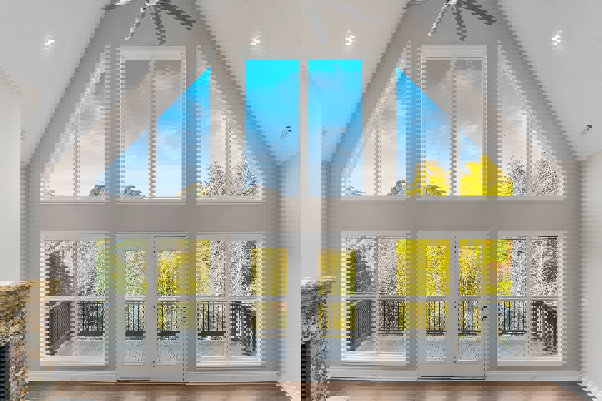 Spacious room with wooden floors, stone fireplace, ceiling fan, large windows showing blue sky, and glass door opening to balcony