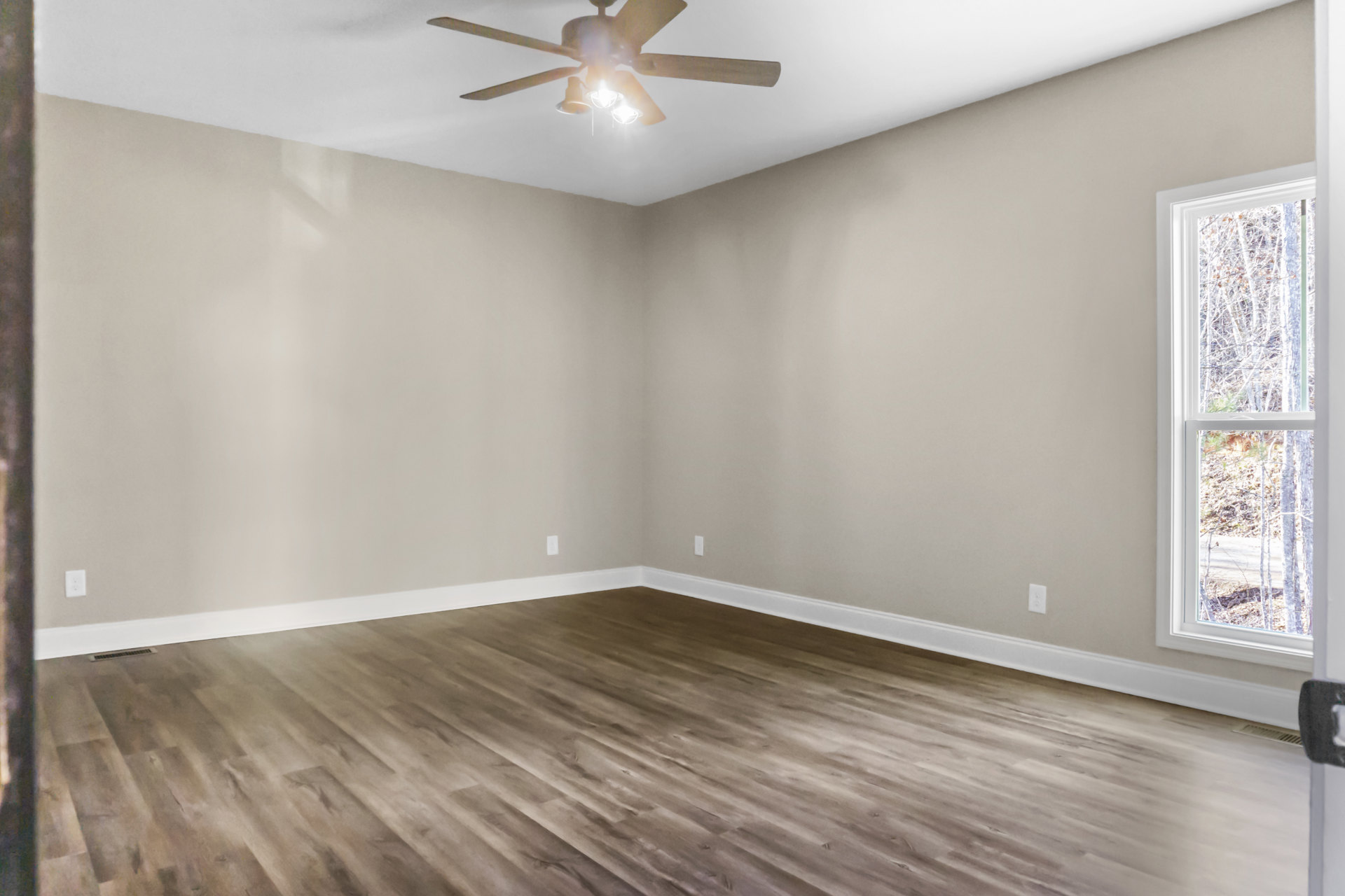 Ceiling fan with illuminated light fixture above wood flooring, white trim, and grey and white walls; window reveals trees outside