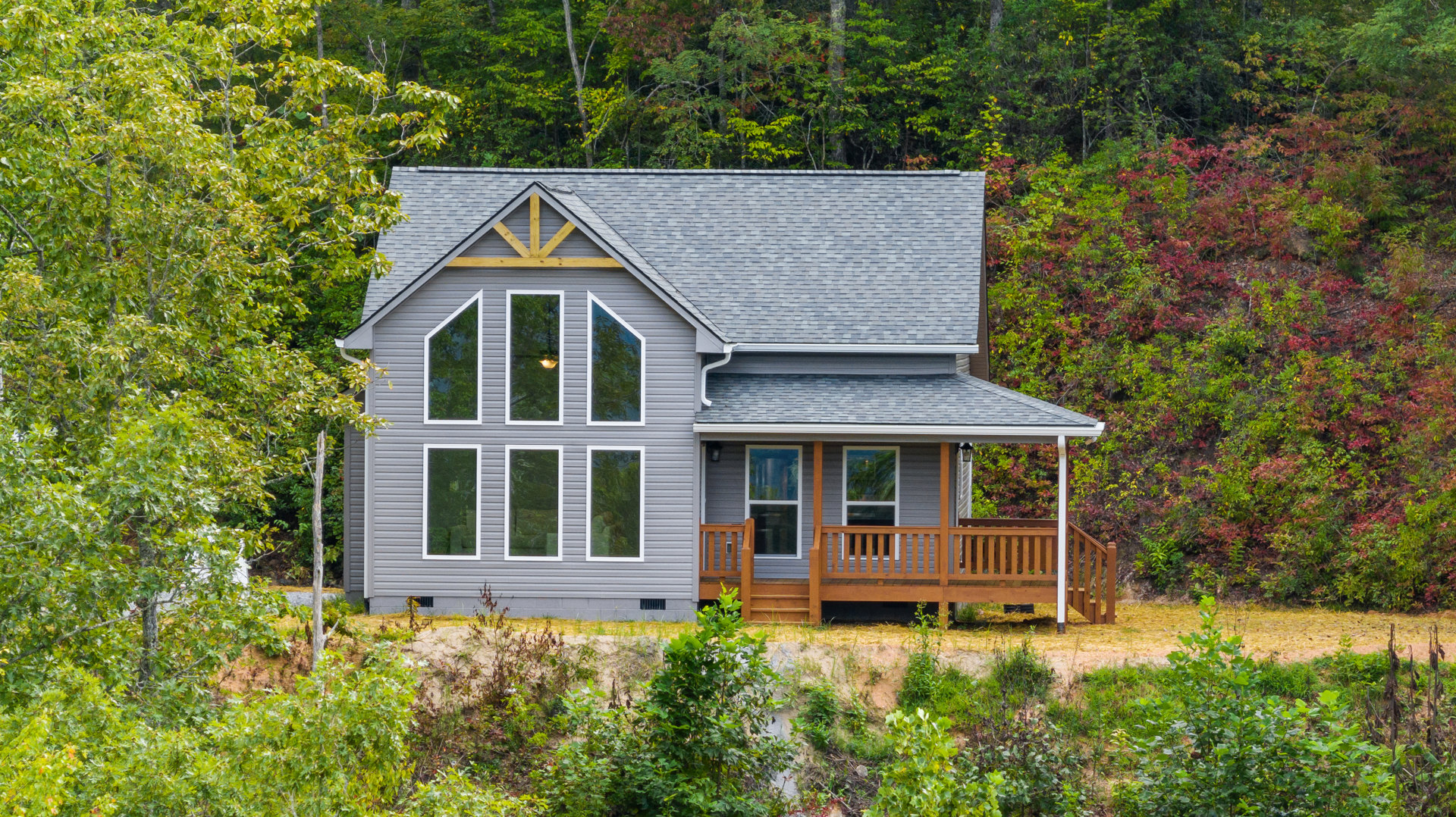 Wooden cottage with wraparound porch and deck, surrounded by tall trees; railing, multiple windows reflecting greenery, warm interior light visible through glass