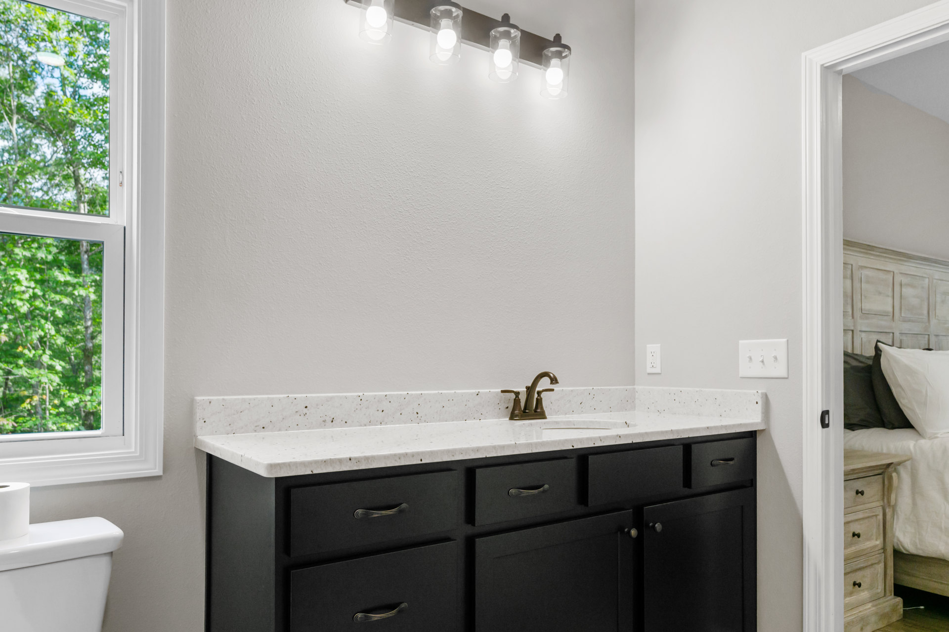 Bathroom with white tile walls, single sink set in a stone countertop, chrome faucet, glass light fixtures above mirror, window showing green trees, toilet with toilet paper roll